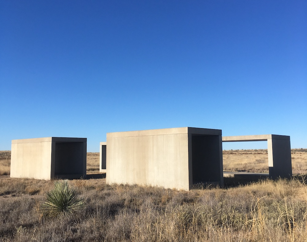 A group of large rectangular concrete forms on the scrubby desert ground with blue sky above